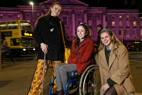 Roísin Hackett, Niamh Ní Hoireabhaird and Laura Beston of Trinity Disability Group, outside a purple TCD style=