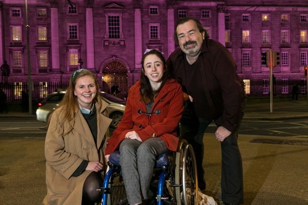 Laura Beston, Niamh Ní Hoireabhaird and disability activist Gary Kearney outside a purple TCD style=